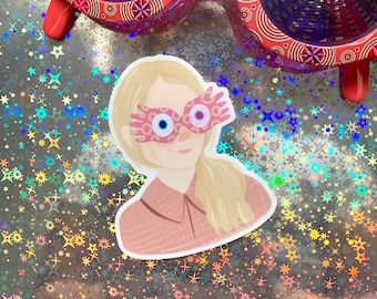 May include: A sticker of a woman with blonde hair wearing pink glasses with a golden design. The glasses have large, round, blue eyes. The woman is wearing a pink shirt. The sticker is on a silver background with a holographic pattern of stars.