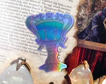 May include: A holographic sticker of a golden goblet with the words "Triwizard" written on it. The sticker is on a page of a book with text about the Triwizard Tournament.
