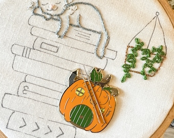 May include: A white cat embroidered on a cream-colored fabric with a stack of books underneath. A small pumpkin house with a green door and a window is attached to the fabric. A hanging plant is embroidered on the right side of the fabric.