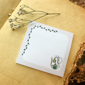 May include: A white notepad with a green vine border and a teapot illustration. The teapot has a green roof and windows with yellow light.