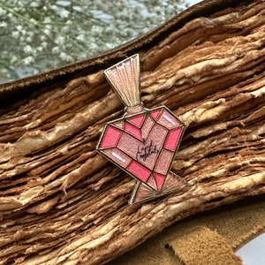 May include: A gold-toned enamel pin shaped like a heart-shaped bottle with a pink and gold design. The bottle has a gold cap and the text "Liquid Crystals" is written on the front.