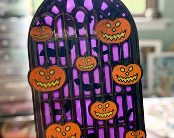 May include: A black metal pin shaped like a stained glass window with a purple background and orange pumpkins with jack-o-lantern faces. There are bats flying around the pumpkins.