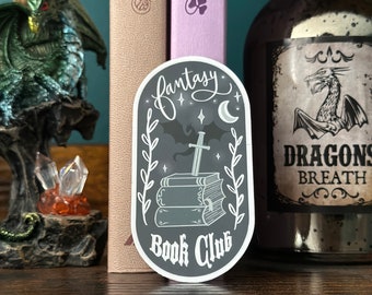May include: A black and white sticker with a fantasy book club theme. The sticker features a stack of books with a sword sticking out of the top. The text "fantasy book club" is written in a decorative font.