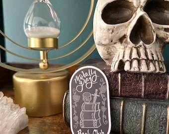May include: A sticker with the text "Morally Grey Book Club" in white script. The sticker features a skull, sword, and books in a dark gray color, with floral accents. The sticker is on a wooden surface.