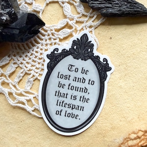 May include: A white ceramic mug with a black handle and the text "But First, Coffee" in a bold, black font. The mug is set against a blurred background, suggesting a cozy setting.