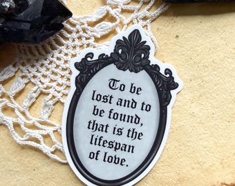 May include: A white ceramic mug with a black handle and the text "But First, Coffee" in a bold, black font. The mug is set against a blurred background, suggesting a cozy setting.