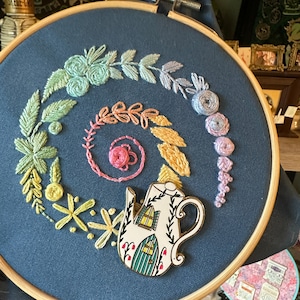 May include: A blue embroidery hoop featuring a colourful floral design and a teapot-shaped pin with a house design. The pin is white with green trim and a yellow window.