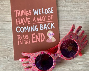 May include: A brown card with the text "Things we lose have a way of coming back to us in the end."  A pink illustration of a woman with pink glasses is on the card.  A pair of pink plastic glasses with a hand-shaped frame and a sparkly design are on a wooden surface.