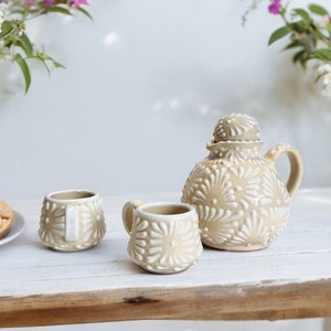 Puede incluir: Un juego de té de cerámica beige con un diseño floral, que incluye una tetera, dos tazas y un plato de galletas. El juego está dispuesto sobre una mesa de madera blanca rústica, con flores en el fondo.