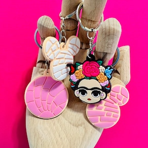 May include: A set of three keychains featuring a pink and white concha, a Frida Kahlo inspired design, and a pink and white Mickey Mouse inspired design.