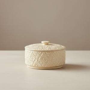 May include: A cream-colored ceramic jar with a lid. The jar and lid are decorated with a repeating floral pattern. The jar is round with a small base and a small knob on the lid.