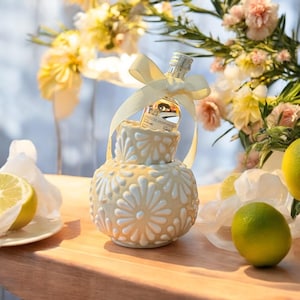 May include: A white ceramic vase with a floral design, holding a small bottle of liquor tied with a white ribbon. The vase is sitting on a wooden surface with lemons nearby.