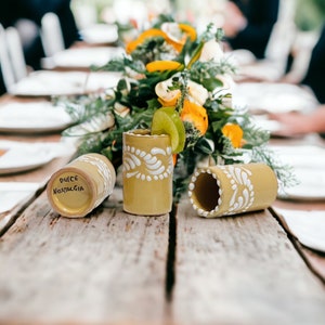 May include: Three yellow ceramic shot glasses with white painted designs. The glasses are on a wooden table with a floral centerpiece in the background. One glass has the words "Dulce Nostalgia" written on the bottom.