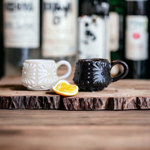 May include: Two small ceramic mugs with a raised floral design. One mug is white and the other is black. The mugs are sitting on a wooden surface with a slice of orange in between them.