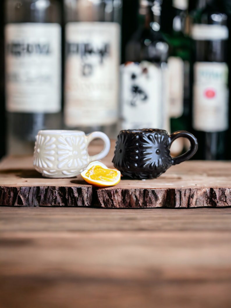 May include: Two small ceramic mugs with a raised floral design. One mug is white and the other is black. The mugs are sitting on a wooden surface with a slice of orange in between them.
