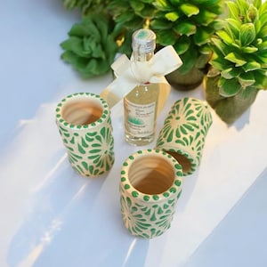 May include: Three small ceramic shot glasses with a green and white floral design. A small bottle of tequila is in the background.