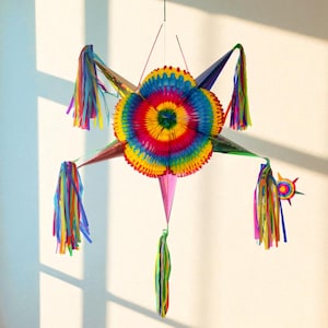 May include: A vibrant, star-shaped piñata in a rainbow of colours. The central flower-like shape has concentric circles of red, orange, yellow, and blue. Long, colourful streamers and a starburst add to the design.