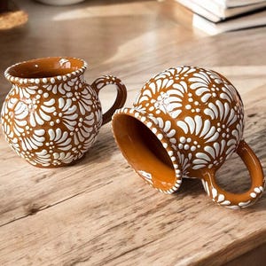 May include: Two brown ceramic mugs with white floral designs. The mugs have a rounded body and a handle.