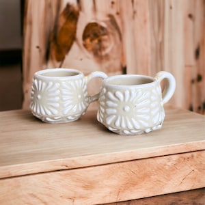 May include: Two white ceramic mugs with a raised floral design. The mugs are sitting on a wooden surface.