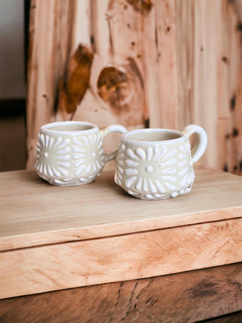 May include: Two white ceramic mugs with a raised floral design. The mugs are sitting on a wooden surface.