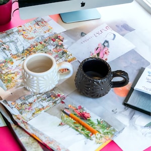 May include: Two ceramic mugs, one white with a raised dot pattern and one black with a raised star pattern, on a table with papers and a pencil.