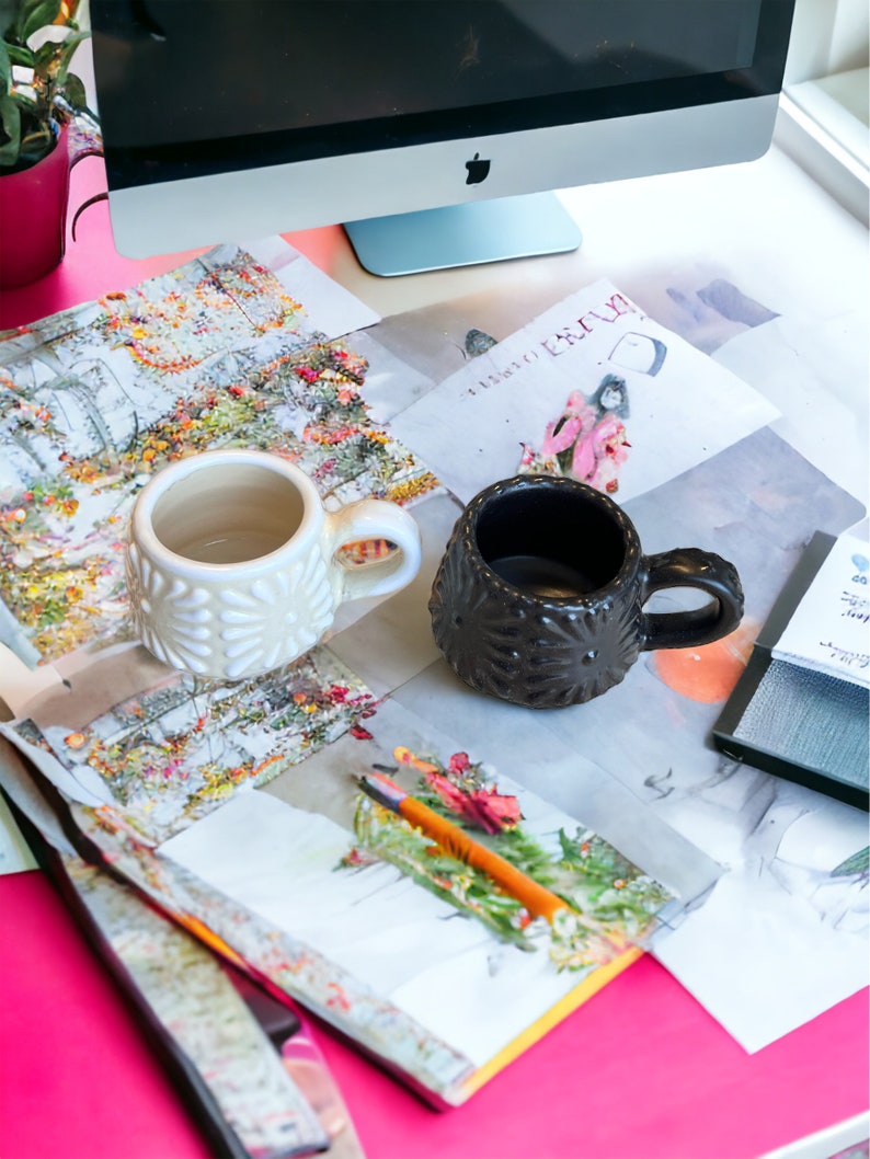 May include: Two ceramic mugs, one white with a raised dot pattern and one black with a raised star pattern, on a table with papers and a pencil.