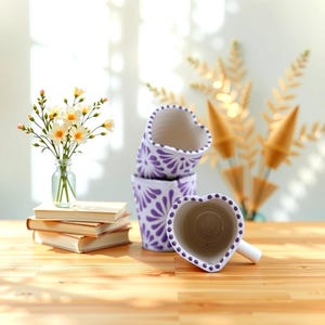 May include: Four purple and white heart-shaped ceramic planters with a floral design. The planters are stacked on top of each other, with the largest one at the bottom and the smallest one at the top. The planters are sitting on a wooden table.