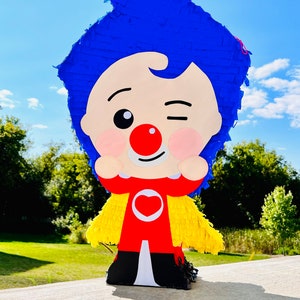 Puede incluir: Una piñata con forma de personaje que presenta una gran cabeza con flecos azules, una nariz roja y una expresión de guiño. La figura viste un traje rojo con un emblema de corazón blanco en el pecho y una capa con flecos amarillos, sobre pies negros.