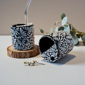 Pode incluir: Dois recipientes de cerâmica em preto e branco com um padrão floral. Um recipiente vertical tem um canudo de vidro transparente. O outro recipiente está de lado. Ambos estão em uma superfície branca com um porta-copos de madeira.