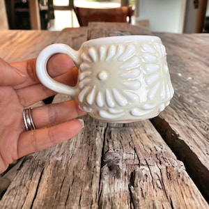 May include: A white ceramic mug with a raised floral design. The mug has a rounded shape and a handle.