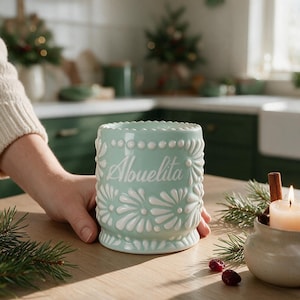 May include: A light mint green ceramic mug featuring a raised white floral pattern and the word Abuelita written in white script on the front. The mug has a scalloped top edge and a textured surface with a glossy finish, designed for holding beverages.