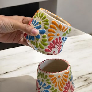May include: Two ceramic flower pots with a white base and colorful floral designs in blue, green, orange, and pink. One pot is held up, revealing the interior, while the other sits below. The pots have a square shape with rounded edges.