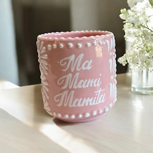 Könnte beinhalten: Eine rosa Keramikvase mit den Worten "Ma Mami Mamita" in weißer Schrift. Die Vase hat eine strukturierte Oberfläche mit weißen Punkt- und Blumendesigns. Die Vase steht auf einer weißen Oberfläche.