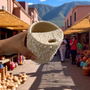 Puede incluir: Jardinera de cerámica color crema con un diseño floral en relieve. La jardinera tiene forma de media luna con un agujero para colgar. El fondo muestra un mercado con edificios y montañas.