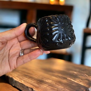 May include: A small black ceramic mug with a textured, floral design. The mug has a rounded shape and a small handle.