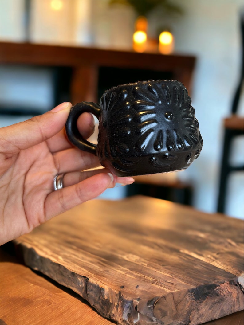 May include: A small black ceramic mug with a textured, floral design. The mug has a rounded shape and a small handle.