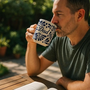 May include: A white ceramic mug with a blue floral design and the word "Dad" in script. The mug is held by a person, who is drinking from it. The mug is on a wooden table, with a book open in front of it.