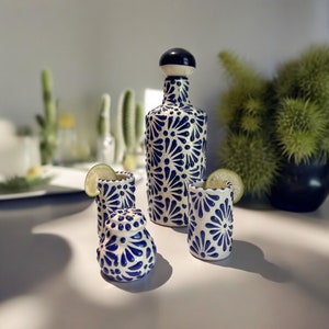 May include: A ceramic tequila set featuring a bottle with a black and white stopper, three shot glasses, and a small container. The set is decorated with a blue floral pattern. Lime wedges are placed on the shot glasses.