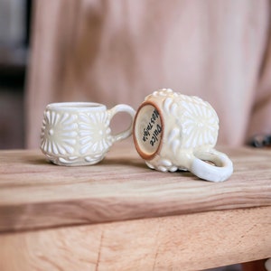 May include: Two white ceramic mugs with a raised floral design. One mug has the words "Juice  Nostalgia" written on it.