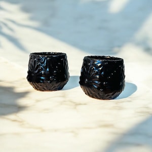 May include: Two black ceramic cups with a textured, floral design. The cups are sitting on a white marble surface.