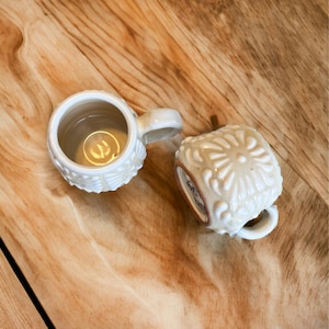 May include: Two white ceramic mugs with embossed floral designs. One mug is filled with a light brown liquid.
