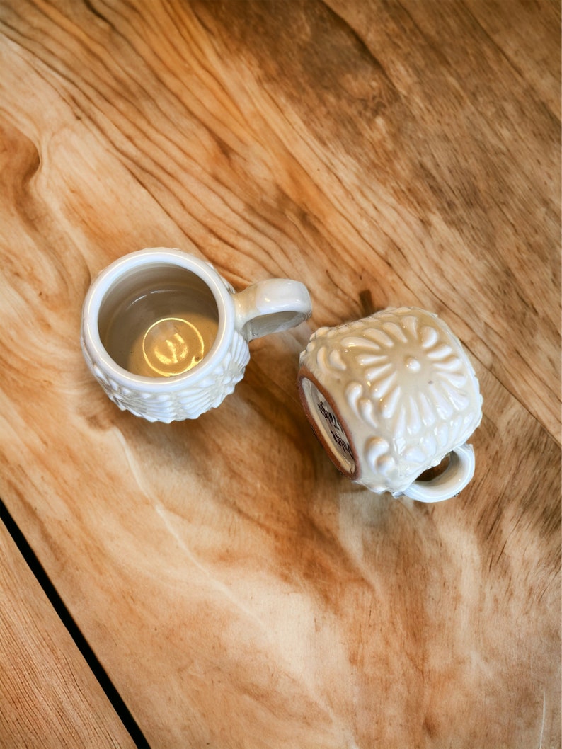 May include: Two white ceramic mugs with embossed floral designs. One mug is filled with a light brown liquid.