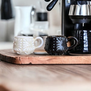 Puede incluir: Dos tazas de cerámica, una blanca con un patrón floral y otra negra con un patrón floral, se encuentran sobre una tabla de cortar de madera. Una máquina de espresso negra está en el fondo.
