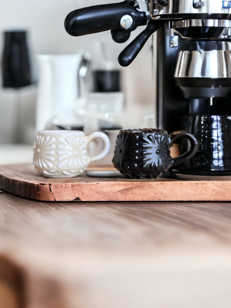 May include: Two ceramic mugs, one white with a floral pattern and one black with a floral pattern, sit on a wooden cutting board. A black espresso machine is in the background.
