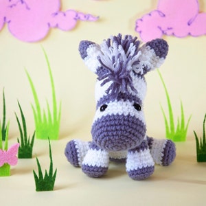 May include: A crocheted zebra toy with white and purple stripes, sitting on a light yellow surface with green felt grass and pink felt clouds in the background.
