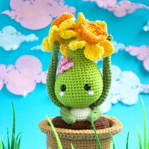 Könnte beinhalten: Ein gehäkeltes Amigurumi-Spielzeug in Form einer Pflanze mit einem grünen Körper, einem braunen Topf und gelben Narzissenblüten. Das Spielzeug hat ein süßes Gesicht mit schwarzen Augen und rosa Wangen. Das Spielzeug sitzt auf einem blauen Hintergrund mit grünem Gras.
