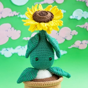 Könnte beinhalten: Eine grüne gehäkelte Pflanze mit einer gelben gehäkelten Sonnenblume oben drauf. Die Pflanze steht in einem braunen gehäkelten Topf. Der Hintergrund ist grün mit rosa und blauen Wolken.