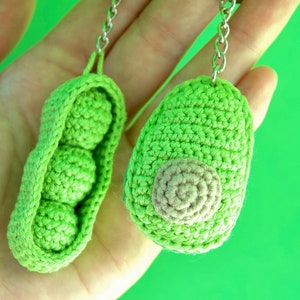 Könnte beinhalten: Zwei gehäkelte Schlüsselanhänger in Form von Erbsenhülsen. Ein Schlüsselanhänger ist offen und zeigt drei grüne Erbsen im Inneren. Der andere Schlüsselanhänger ist geschlossen und hat ein braunes Häkeldetail an der Seite.