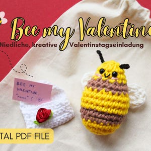 Könnte beinhalten: Eine gehäkelte Biene mit braunen und gelben Streifen und eine weiße gehäkelte Tasche mit einer rosa Rose und einem Zettel mit der Aufschrift "Bee my Valentine." Der Hintergrund ist eine rote Oberfläche.