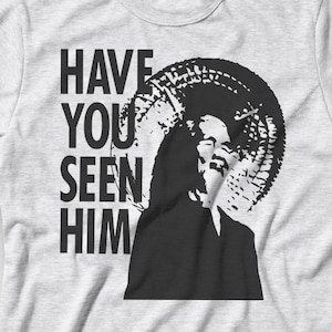 May include: A light gray t-shirt featuring a black and white graphic of a person with a halo-like design. The text "HAVE YOU SEEN HIM" is printed vertically on the left side of the graphic.